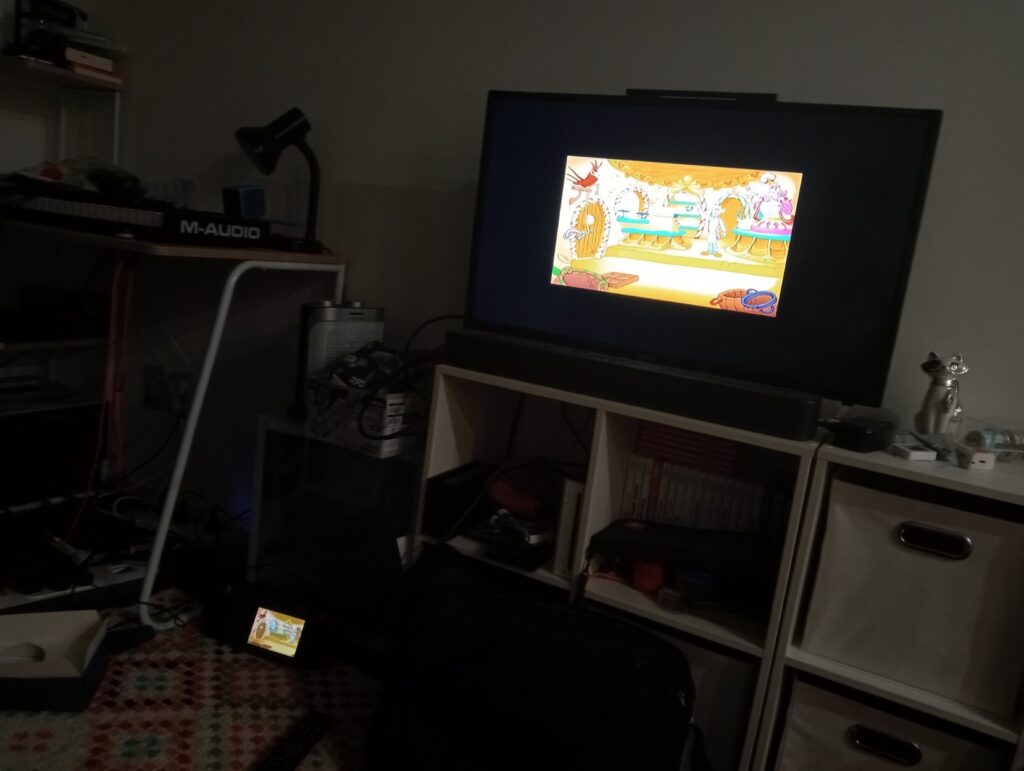 Photograph of a steam deck plugged into a television. You can see a low resolution colorful shovelware game on the screen. 